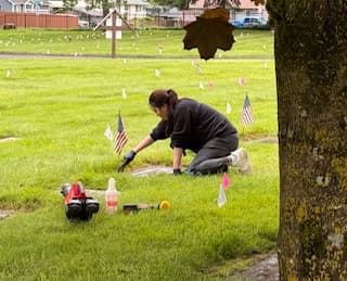 Woman kneels in a grassy cemetery cleaning a flat grave marker near small American flags.