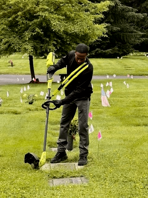 A man trims grass around a grave marker in a cemetery with a string trimmer.
